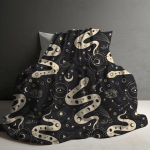 May include: A black and cream throw blanket with a repeating pattern of a snake, stars, moons, and eyes. The snake is winding its way through the pattern, and the stars and moons are scattered around it.