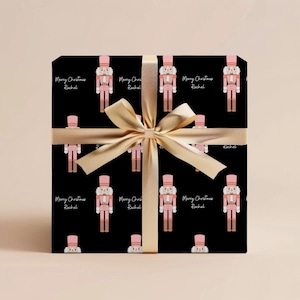 May include: Black wrapping paper with a repeating pattern of pink nutcracker illustrations and the text "Merry Christmas Rachel". The paper is wrapped around a gift box with a gold ribbon.