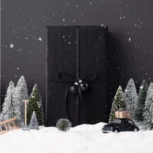 May include: A black wrapped gift with a black ribbon and ornaments, set in a winter scene with miniature snow-covered trees and a toy car carrying packages. Snowflakes fall against a dark background.