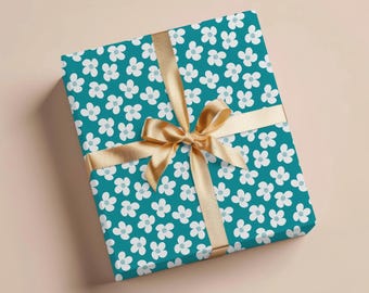 May include: A wrapped gift box featuring teal floral patterned paper with white and light blue flowers. The box is tied with a gold ribbon and bow, creating a festive presentation. The background is a solid, neutral color.