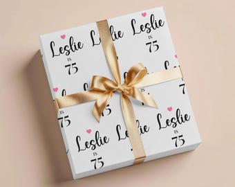 May include: A wrapped gift box with a white background and a repeating pattern of the name "Leslie", the number "75", and pink hearts. A gold ribbon and bow are tied around the box.
