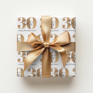 Pode incluir: Um presente embrulhado com papel branco com texto dourado "30" e "Thirty". As palavras "Happy Birthday Sarah" tamb&eacute;m est&atilde;o impressas no papel. Uma fita dourada &eacute; amarrada em um la&ccedil;o ao redor do presente.