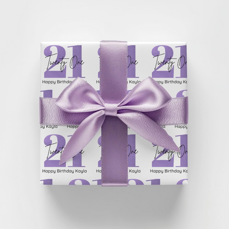 Pode incluir: Um presente embrulhado com fundo branco e um padr&atilde;o repetido do n&uacute;mero "21" e as palavras "Twenty One". O presente &eacute; amarrado com uma fita e la&ccedil;o de cetim lavanda. O texto "Happy Birthday Kayla" tamb&eacute;m &eacute; impresso no papel de embrulho.