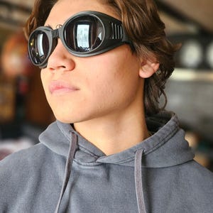 Könnte beinhalten: Schwarze, runde Schutzbrille mit einem dicken, verstellbaren Riemen. Die Gläser sind dunkel und reflektieren die Umgebung. Die Brille wird über den Augen getragen. Die Person trägt einen grauen Kapuzenpullover.