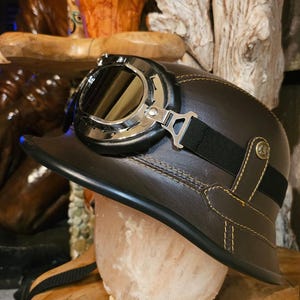 Puede incluir: Un casco de moto de cuero marrón con gafas con montura plateada. El casco tiene un borde negro y costuras amarillas. Una correa negra asegura las gafas. El casco está diseñado al estilo vintage, que recuerda al equipo de moto clásico.