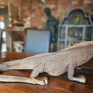 May include: A detailed wooden sculpture of a Komodo dragon, with textured scales and a long tail, rests on a polished wooden surface. The reptile is carved in a naturalistic pose, showcasing its legs and head. The sculpture is a decorative item.