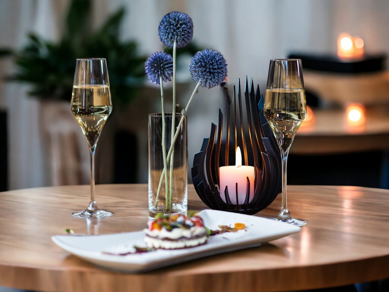 May include: A table setting with two glasses of white wine, a plate with a dessert, a vase with blue flowers, and a candle holder with a lit candle.