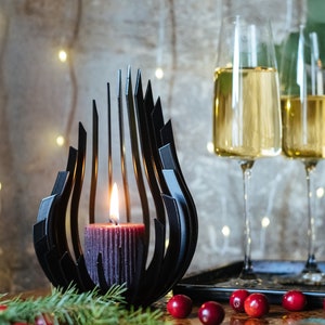 May include: A black metal candle holder shaped like a flame with a dark red candle inside. Two champagne flutes filled with a golden liquid are in the background.