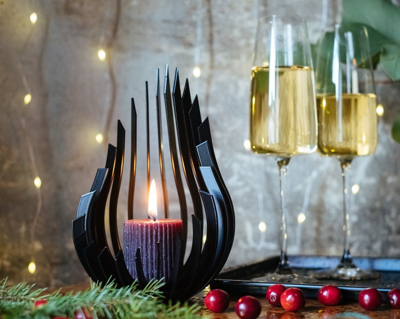 May include: A black metal candle holder shaped like a flame with a dark red candle inside. Two champagne flutes filled with a golden liquid are in the background.