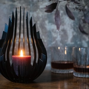 May include: A black metal candle holder with a unique, abstract design. A lit candle sits inside the holder, casting a warm glow. Two clear glass tumblers with dark liquid are in the background.
