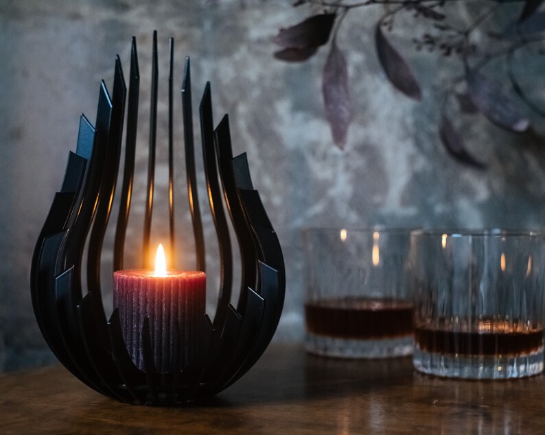 May include: A black metal candle holder with a unique, abstract design. A lit candle sits inside the holder, casting a warm glow. Two clear glass tumblers with dark liquid are in the background.