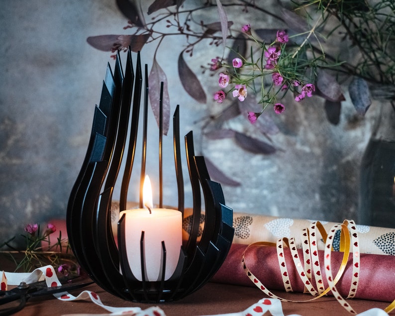 May include: A black metal candle holder shaped like a flame with a white candle inside. The candle holder is sitting on a table with pink and white ribbon and a pair of scissors.