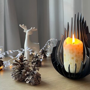 May include: A black metal candle holder with a modern design holds a white candle with gold stars and circles. The candle is lit and the flame is visible. The candle holder is on a wooden table with a white ceramic deer figurine and pine cones.