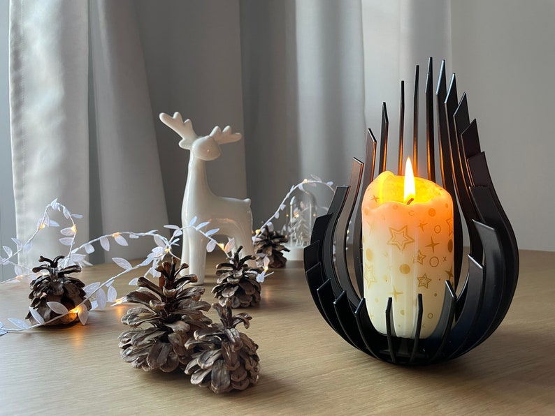 May include: A black metal candle holder with a modern design holds a white candle with gold stars and circles. The candle is lit and the flame is visible. The candle holder is on a wooden table with a white ceramic deer figurine and pine cones.