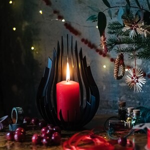 May include: A black metal candle holder shaped like a flame with a red candle burning inside. The candle holder is on a wooden table with red berries and red ribbon.