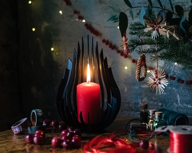 May include: A black metal candle holder shaped like a flame with a red candle burning inside. The candle holder is on a wooden table with red berries and red ribbon.