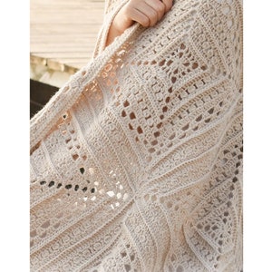 May include: A cream-coloured, open-weave crochet shawl with a geometric pattern. The shawl features square and diamond shapes with intricate detailing. The yarn appears to be a soft, natural fibre, creating a delicate and textured look. The shawl is draped over a person's arm.