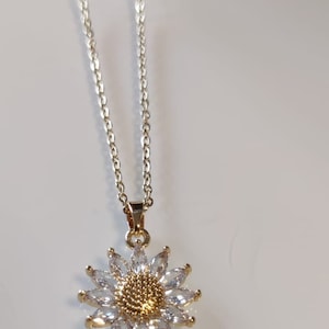 Puede incluir: Un collar de cadena dorada con un colgante de girasol. El colgante está hecho de diamantes de imitación transparentes y tiene un centro dorado.