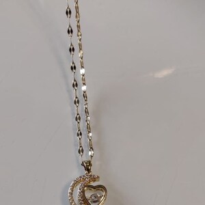 Puede incluir: Un collar de oro con un colgante de media luna que presenta un corazón con un cristal transparente en el centro. El colgante está adornado con pequeños cristales transparentes.