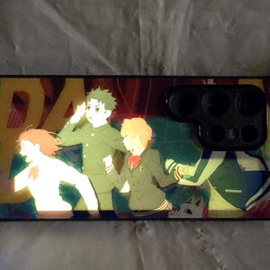Puede incluir: Una funda para teléfono con un diseño de anime colorido con cuatro personajes. La funda tiene un borde negro y un recorte para la cámara con múltiples lentes.