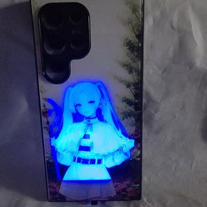 Puede incluir: Una funda de teléfono negra con un diseño de personaje de anime azul y blanco. El personaje es una joven con el pelo largo azul y un vestido blanco. La funda tiene una parte trasera transparente y un marco negro.