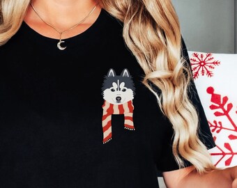 May include: A black t-shirt with a graphic of a husky wearing a red and white scarf. The person wearing the shirt is sitting on a white and red blanket.