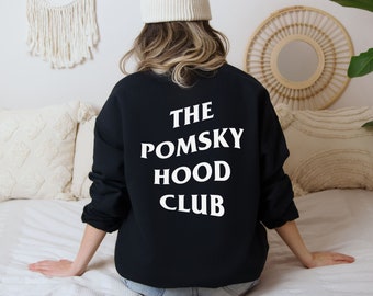 May include: Black sweatshirt with the text "THE POMSKY HOOD CLUB" in white, centered on the back. The sweatshirt is a solid black color. The person is wearing a white beanie.