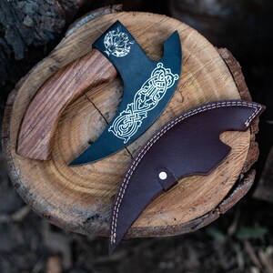 May include: A black axe with a wooden handle and a leather sheath. The axe has a decorative design on the blade and the sheath has a stitched edge.
