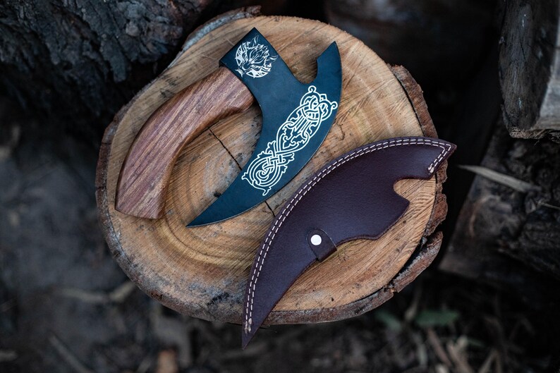 May include: A black axe with a wooden handle and a leather sheath. The axe has a decorative design on the blade and the sheath has a stitched edge.