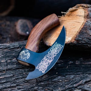 May include: A black axe with a wooden handle and intricate etched designs. The axe is resting on a piece of wood.