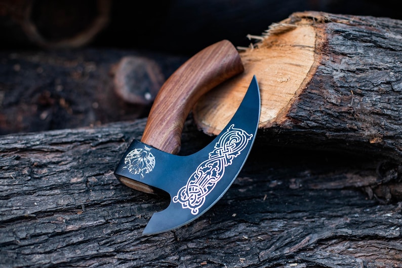 May include: A black axe with a wooden handle and intricate etched designs. The axe is resting on a piece of wood.
