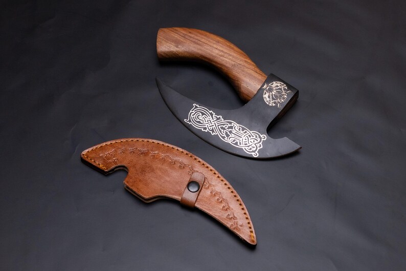 May include: A black and silver axe with a wooden handle and a leather sheath. The axe has an intricate design on the blade.