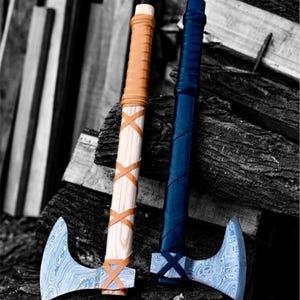 Könnte beinhalten: Zwei Wikingeräxte mit Holzstielen, die in Leder gewickelt sind. Die Axt links hat eine braune Lederwicklung und die Axt rechts eine schwarze Lederwicklung. Beide Äxte haben eine Damastmesserklinge.