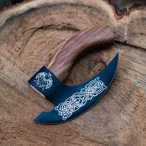 May include: A black axe with a wooden handle. The axe head has a decorative design etched into it. The axe is lying on a wooden surface.