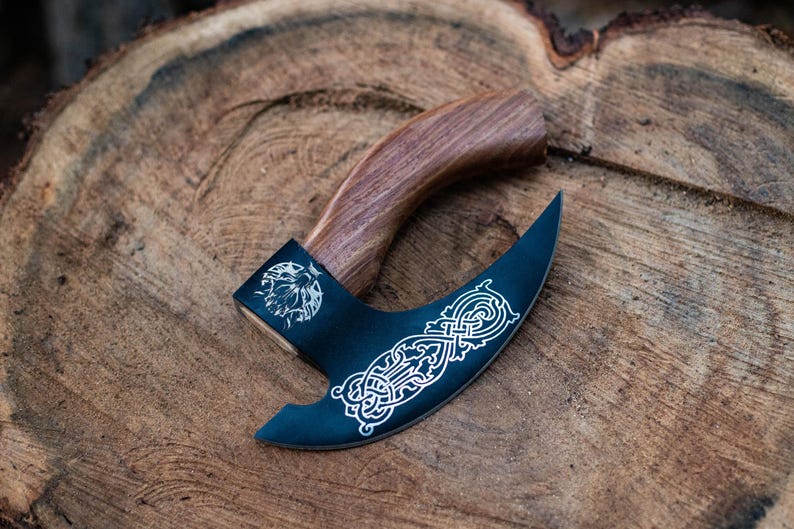 May include: A black axe with a wooden handle. The axe head has a decorative design etched into it. The axe is lying on a wooden surface.