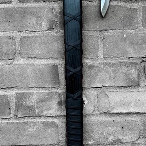 Könnte beinhalten: Eine schwarze und silberne Axt mit einem Holzgriff, der mit schwarzem Leder umwickelt ist. Der Axtkopf ist silberfarben und der Griff ist mit schwarzen Lederriemen befestigt. Die Axt wird vor einer grauen Backsteinmauer ausgestellt.