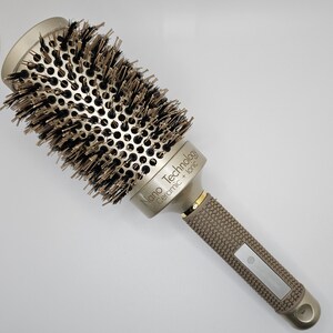May include: A round, gold-toned hairbrush with black and brown bristles. The brush features a textured, brown handle and the text "Nano Technology Ceramic + Ionic". The brush is designed for styling and smoothing hair.
