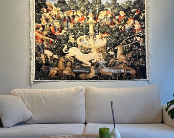 May include: A tapestry depicting a medieval hunting scene with figures, animals, and a fountain. The artwork features a unicorn, deer, and other creatures in a lush forest setting. The tapestry is rectangular with a fringed edge and is mounted on a wall above a sofa.