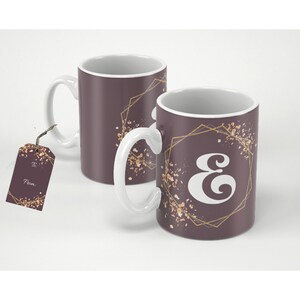 May include: Two white ceramic mugs with a brown background and gold geometric designs. The mugs feature a white letter "E" in a stylised font. One mug has a gift tag that says "To" and "From".