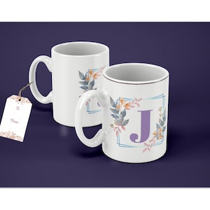 Pode incluir: Duas canecas de cerâmica branca com alças. A caneca à direita tem um desenho floral com uma letra J roxa no centro. A caneca à esquerda tem um desenho semelhante, mas sem a letra.