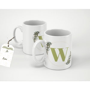 May include: Two white ceramic mugs featuring a green letter "W" design, encircled by a floral wreath. A gift tag with "To:" and "From:" is attached to one mug. Perfect for hot beverages.
