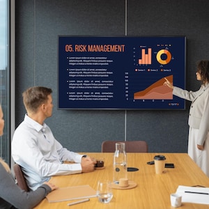 May include: A business presentation in a modern office setting. A large screen displays a "Risk Management" chart with graphs and data. Three people are seated at a wooden table, with one person pointing at the screen. A window and coffee cups are also visible.