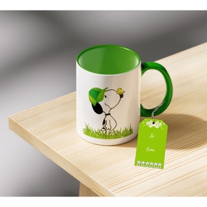 Puede incluir: Una taza de cerámica blanca con interior y asa verdes presenta al personaje de dibujos animados Snoopy con gorra verde y pájaro amarillo. Una etiqueta de regalo verde con detalles florales blancos y texto "Para: De:" cuelga cerca.