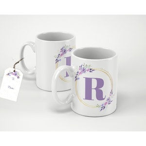 Pode incluir: Duas canecas de cerâmica branca com um design de coroa de flores e a letra "R" em lavanda. Uma etiqueta de presente está anexada a uma caneca. As canecas têm uma borda circular dourada com flores e folhas de lavanda.