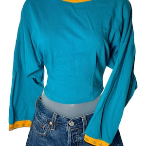 May include: Teal long-sleeve crop top with a yellow and black trim on the neckline and cuffs. The top is paired with a denim skirt. The top has a crew neck and long sleeves.
