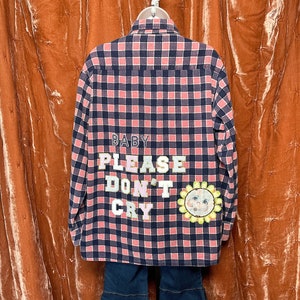 May include: A pink and black plaid flannel shirt with the text "BABY PLEASE DON'T CRY" in colorful letters and a yellow sunflower with a cartoon face on the back.