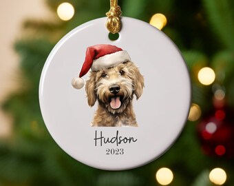 May include: White ceramic Christmas ornament featuring a watercolor illustration of a dog wearing a Santa hat. The dog's name, "Hudson", and the year "2023" are printed below the image. A gold ribbon is attached for hanging.