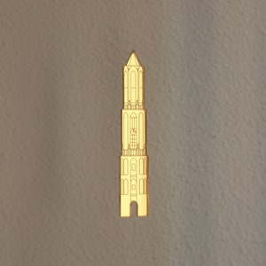 May include: A gold-coloured metal pin shaped like a tall tower with a clock face and multiple windows. The pin is a simple outline design.