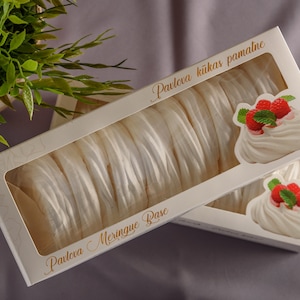 Może przedstawiać: Białe pudełko z przezroczystym okienkiem, w którym widać stos białych bezy. Pudełko jest oznaczone "Pavlova Meringue Base" i "Pavlova kũkas pamatne".