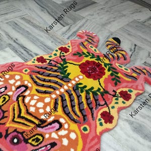 May include: A colorful rug shaped like a tiger with pink, yellow, orange, black, and green accents. The rug has a floral design with a large pink flower in the center. The rug is made of wool and is hand-knotted. The rug is made by Karsten Rugs.
